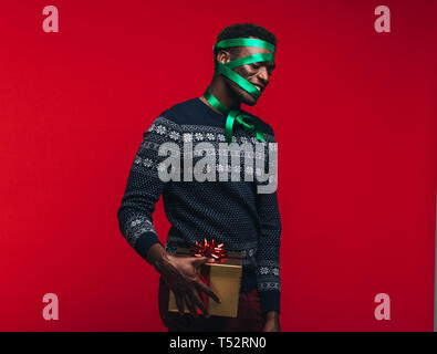 Afrikanischer Mann mit Satinband auf seinem Gesicht Holding ein Geschenk Box verpackt. Mann mit einem Geschenk auf rotem Hintergrund. Stockfoto