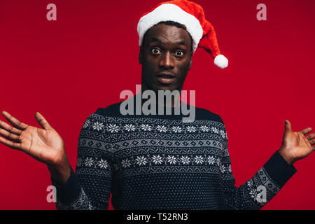 Afrikanische amerikanische Mann mit Santa hat über den roten Hintergrund überrascht. Überrascht leer Kerl auf Kamera. Stockfoto