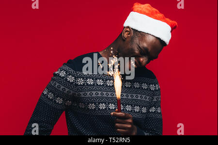 Fröhlicher junger afrikanischer Mann mit Wunderkerzen auf rotem Hintergrund. Jungen Kerl trägt einen santa hut mit Feuerwerk. Stockfoto