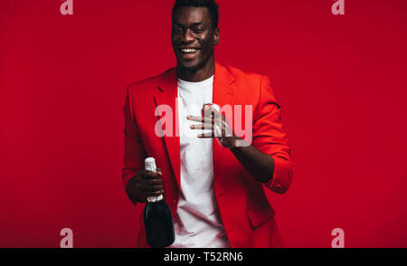 Fröhlicher junger afrikanischer Mann mit Champagner Flasche und Glas. Schöner Mann im roten Mantel mit Champagner und Glas gegen den roten Hintergrund. Stockfoto