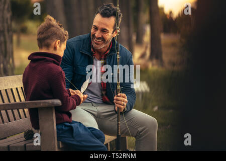 Freundliche Vater und Sohn sitzen draußen auf einer Bank mit einer Angelrute. Lächelnd Mann im Gespräch mit seinem Sohn eine Angelrute. Stockfoto