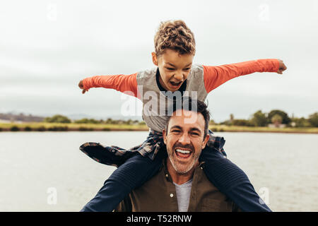 Junge sitzt auf den Schultern seines Vaters und Spielen. Vater und Sohn Spaß Ausgabe Zeit zusammen in der Nähe von einem See. Stockfoto