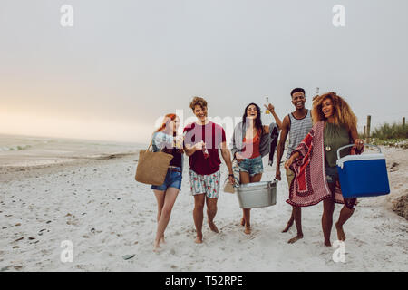 Gruppe von jungen Menschen zu Fuß auf den Strand, eine Kühlbox und Getränke Whirlpool. Junge Männer und eine Frau am Meer. Stockfoto