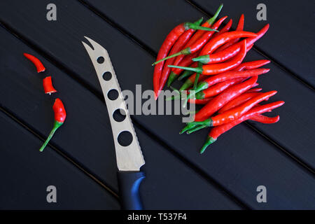 In Scheiben geschnitten und Ganze rote Chilischoten neben Gemüse und Käse Messer liegen auf einem schwarzen Holz- Oberfläche. Tageslicht Stockfoto