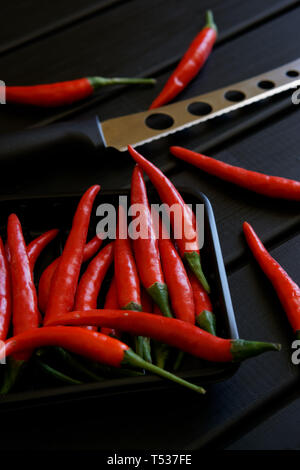 Rote Chili Schoten und ein Messer für Käse und Gemüse liegen auf einem schwarzen Holz- Oberfläche. Tageslicht. Bild vertikal. Close-up Stockfoto