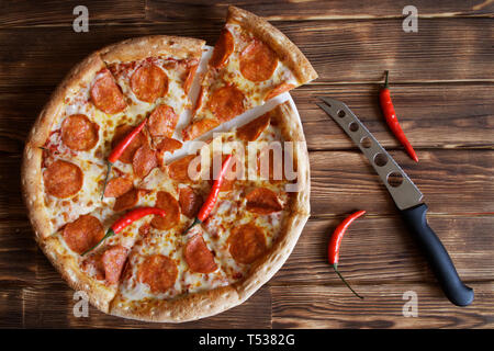 Hausgemachte Pizza mit Salami und red chili peppers liegt neben einem Käse Messer auf einem natürlichen Holz- Oberfläche aus Kiefern- platten. Schneiden Sie ein Stück. Da Stockfoto