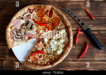 Sortierte Pizza, getrocknete Chilischoten und Käse Messer liegen auf einem natürlichen Holz- Oberfläche von Pine Planken. Schneiden Sie ein Stück. Tageslicht. Stockfoto