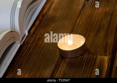 Eine brennende Kerze steht auf Holzplanken neben einem offenen Weißen Buch der Religion und des Glaubens. Close-up.