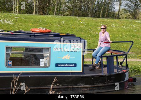 Vorderansicht der isolierten lächelnden weiblichen Dame am hinteren Ende des Schmalboots, das Boot steuern, Reisen in gemächlichem Tempo UK Kanal im Frühlingssonne. Stockfoto