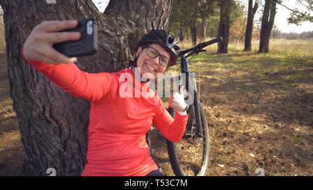 Junge kaukasier Sportlerin touristische Radfahrer verwendet hand smart phone Foto von sich selfie sitzen in der Nähe von Tree In den herbstlichen Wald außerhalb der Stadt Stockfoto