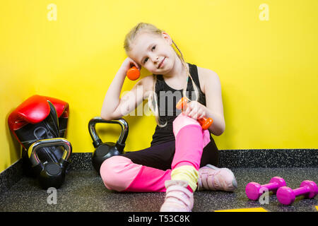 Thema Sport und Gesundheit der Kinder. Kleine lustige Kind kaukasische Mädchen mit Zöpfen, sitzt auf dem Boden ruht in der Turnhalle. Sportler Hantel Ausrüstungen für Stockfoto
