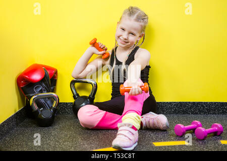 Thema Sport und Gesundheit der Kinder. Kleine lustige Kind kaukasische Mädchen mit Zöpfen, sitzt auf dem Boden ruht in der Turnhalle. Sportler Hantel Ausrüstungen für Stockfoto