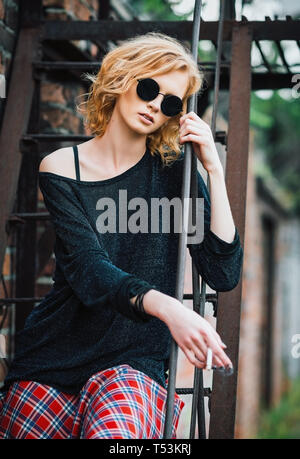 Schöne junge Grunge (Rock) Mädchen mit karierten Rock, Pullover und Brille sitzt auf der Treppe und hält Zigarette Stockfoto