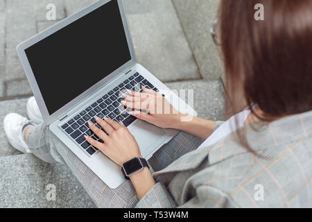 Junge Frau sitzt auf der Straße eingeben Nachricht auf Laptop zurück Ansicht schließen Stockfoto