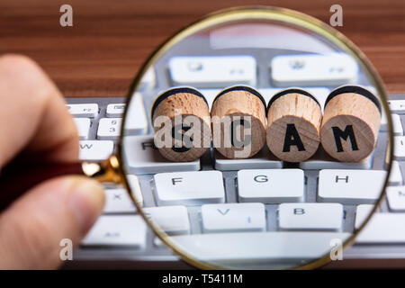 Die menschliche Hand mit Lupe über Holz Kork mit Scam Text auf weißem Tastatur Stockfoto