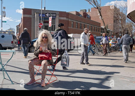 Eine Straße Szene mit einer sitzenden Frau mit langen blonden Haaren Textnachrichten auf Ihr Mobiltelefon. In Jackson Heights, Queens, New York City. Stockfoto