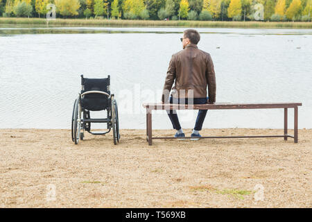 Ein junger Mann sitzt auf einer Bank am See, neben seinem Rollstuhl. Stockfoto