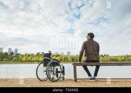 Ein junger Mann sitzt auf einer Bank am See, neben seinem Rollstuhl. Stockfoto