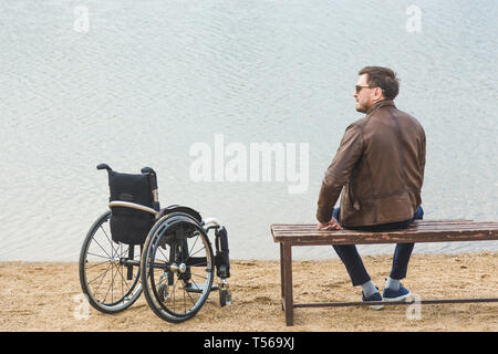 Ein junger Mann sitzt auf einer Bank am See, neben seinem Rollstuhl. Stockfoto
