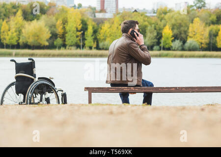 Ein junger Mann sitzt auf einer Bank am See, neben seinem Rollstuhl. Stockfoto