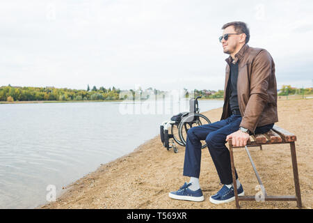 Ein junger Mann sitzt auf einer Bank am See, neben seinem Rollstuhl. Stockfoto