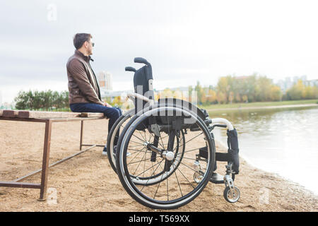 Ein junger Mann sitzt auf einer Bank am See, neben seinem Rollstuhl. Stockfoto