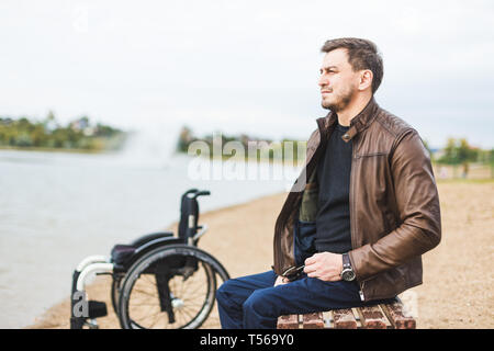 Ein junger Mann sitzt auf einer Bank am See, neben seinem Rollstuhl. Stockfoto