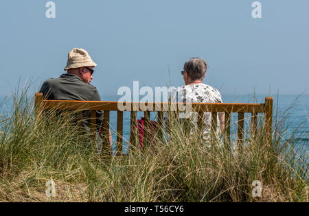 Zwei ältere Menschen oder ein älteres Rentnerehepaar sitzt auf einer Holzbank am Meer im Sommer. Stockfoto