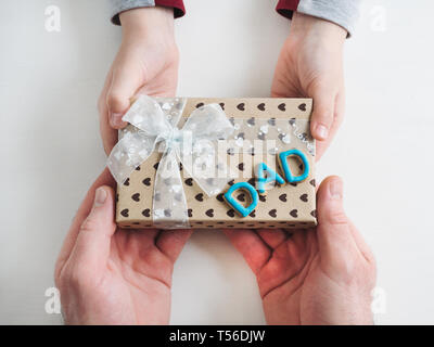 Die Hände des Kindes und die Hände des erwachsenen Mannes, schönen Geschenkkarton, Farbband und glasierte Plätzchen auf einem weißen, Holz- Hintergrund. Ansicht von oben, close-up. Vorbereitung für Th Stockfoto