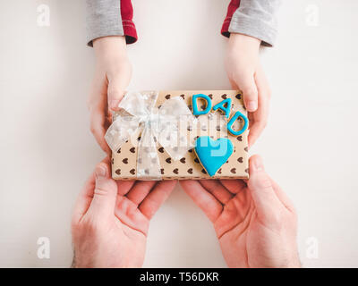 Die Hände des Kindes und die Hände des erwachsenen Mannes, schönen Geschenkkarton, Farbband und glasierte Plätzchen auf einem weißen, Holz- Hintergrund. Ansicht von oben, close-up. Vorbereitung für Th Stockfoto