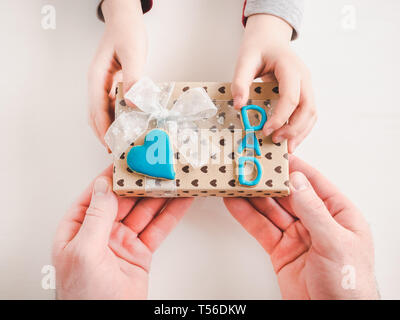 Die Hände des Kindes und die Hände des erwachsenen Mannes, schönen Geschenkkarton, Farbband und glasierte Plätzchen auf einem weißen, Holz- Hintergrund. Ansicht von oben, close-up. Vorbereitung für Th Stockfoto