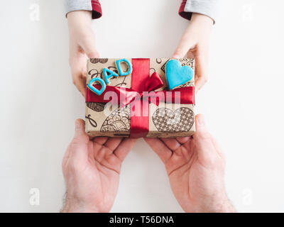 Die Hände des Kindes und die Hände des erwachsenen Mannes, schönen Geschenkkarton, Farbband und glasierte Plätzchen auf einem weißen, Holz- Hintergrund. Ansicht von oben, close-up. Vorbereitung für Th Stockfoto