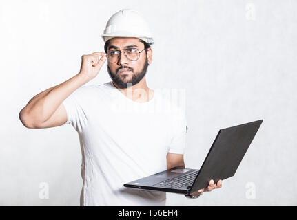 Junge Baumeister Ingenieur in Gläsern und einer weißen Schutzhelm, steht mit dem offenen Laptop. Stockfoto