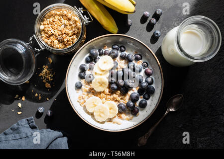 Gesundes Frühstück Hafer Müsli mit Milch, Heidelbeere und Banane auf Schwarz konkreten Hintergrund. Table Top Aussicht, grelles Licht Foodfotografie Stockfoto