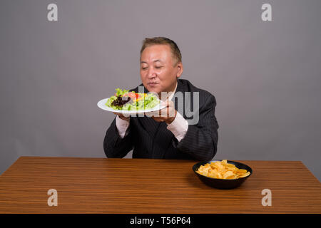 Reifen asiatische Geschäftsmann sitzen mit hölzernen Tisch gegen Grau Stockfoto