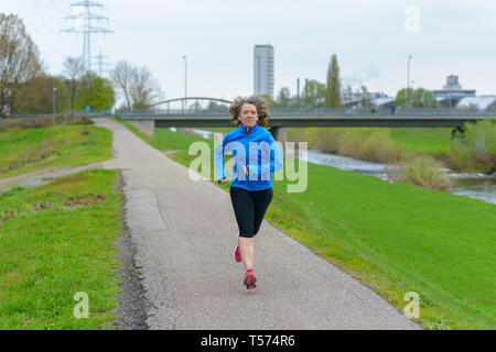 Fit athletische Frau mittleren Alters joggen auf einer Straße neben einem Kanal nähert sich die Kamera. Vorne in voller Länge Porträt mit Kopie Raum Stockfoto