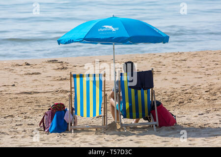 Bournemouth, Dorset, Großbritannien. 22 Apr, 2019. UK Wetter: Nach einem dunstigen Start das herrliche Wetter mit warmen und sonnigen Wetter fort, als beachgoers Kopf zum Meer die Wärme und Sonnenschein in Bournemouth Strände am Ostermontag zu genießen, bevor das Wetter ändert sich und die Rückkehr zur Arbeit. Zwei Liegestühle mit blauen Sonnenschirm mit Mann sitzt in einem Liegestuhl. Credit: Carolyn Jenkins/Alamy leben Nachrichten Stockfoto