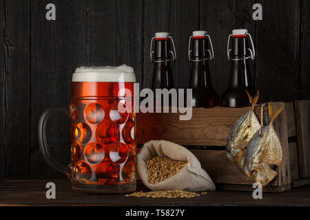 Glas Bier auf Tisch mit Holzkiste voller Flaschen, Beutel mit Bier, Gerste und salzig getrockneten Fisch Stockfoto