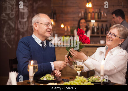 Schöne alte Dame bewundern Sie die Blumen, die ihr Mann ihr gegeben hat. Paar in den 60er Jahren essen. Frischen Weintrauben. Glas Wein. Stockfoto