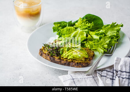 Avocado Toast mit Pesto, Sprossen und Salat auf einem weißen Teller und Eis Kaffee. Stockfoto