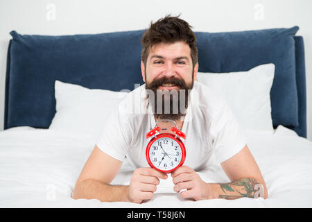 Warum sollten Sie früh aufstehen jeden Morgen. Nutzen für die Gesundheit der Früh aufstehen. Aufwachen frühzeitig, mehr Zeit für die Vorbereitung und zeitgerecht erfolgen. Hipster bärtiger Mann im Bett lagen mit Wecker. Zeit aufzuwachen. Stockfoto