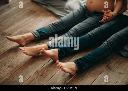 Ein Mann und seine schwangere Frau sitzen auf dem Holzboden und Holg Hände. Stockfoto