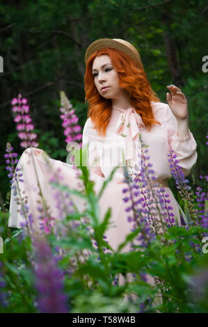 Eine schöne rothaarige Frau steht und stellt in einem Feld von lila Lupinen und geniesst die Aussicht im Frühjahr Stockfoto