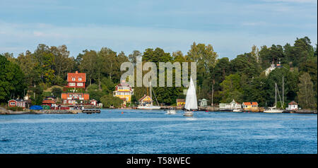 Yachten und Sportboote in einer Bucht, die von einer Ansiedlung von traditionellen bunten Holzhäusern auf Skarpö in den Stockholmer Schären. Stockfoto