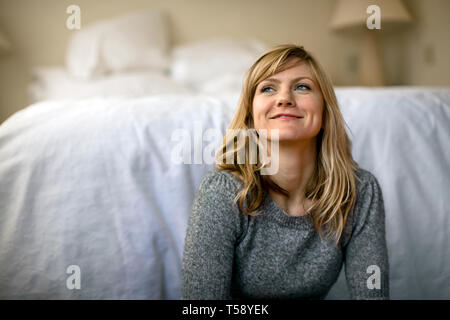 Junge Frau lächelt glücklich in der Ferne, als sie sitzt und Tagträume auf dem Fußboden ihres Schlafzimmers. Stockfoto