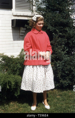 1960er Jahre, USA, hübsche Blondine Jugendmädchen außerhalb in einem Garten für ein Bild tragen modische junge Kleidung, eine Blume gemusterten Rock und Kammgarn rosa-rot oben, mit Stylistin Haarnetz. Stockfoto