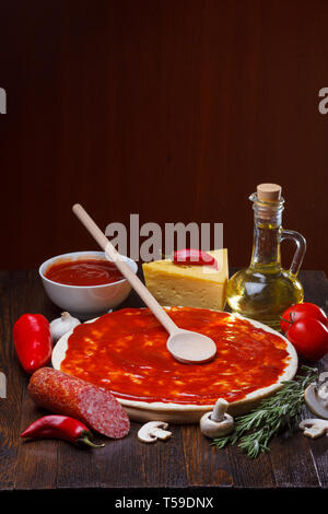 Pizza Teig mit Tomatensauce während der Vorbereitung, mit einer Menge von Zutaten auf hölzernen Tisch Stockfoto
