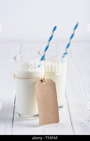 Zwei Gläser Milch mit blauen abgestreift Papier, Stroh und leeres Papier tag am Seil auf weiße Holztisch still life Stockfoto