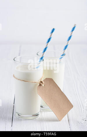 Zwei Gläser Milch mit blauen abgestreift Papier, Stroh und leeres Papier tag am Seil auf weiße Holztisch Stockfoto