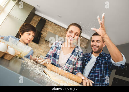 Mutter, Vater und Sohn zu Hause stehend am Tisch in der Küche zusammen Papa spielen mit Mehl lächelt fröhlich während Mama Rollenteig konzentriert Stockfoto